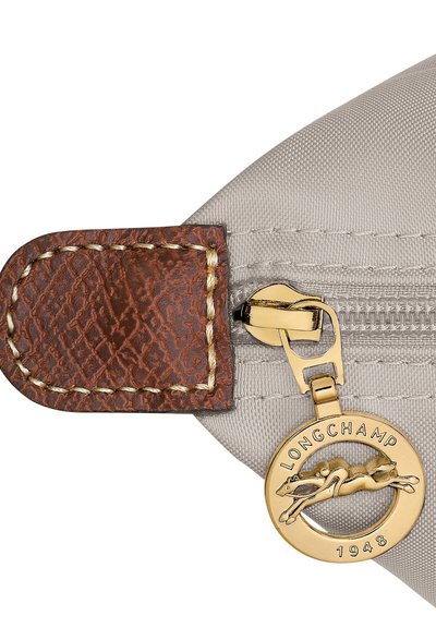 Ecke einer grauen Nylon-Tasche mit einem braunen Leder-Reißverschlussanhänger, der eine strukturierte Oberfläche, goldene Beschläge und einen runden Anhänger mit der Aufschrift "LONGCHAMP 1948" zeigt.