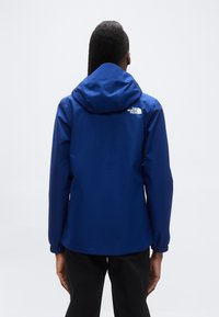 Μπλε αδιάβροχο μπουφάν με κουκούλα, ελαστικά μανίκια και λευκό λογότυπο στην πλάτη. Λείο υφή, σχεδιασμένο για προστασία από τον καιρό.