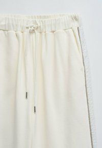 Pantalons de survêtement couleur crème avec une taille élastique, des liens à cordon, et une bande latérale texturée en gris clair et accents argentés.
