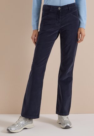 Femme portant un pantalon en velours côtelé bleu foncé à jambes droites et des baskets argentées, debout devant un fond beige.
