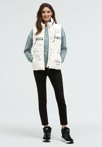 Weißer gepolsterter Westen mit grafischem Textdesign, kombiniert mit einem hellblauen Sweatshirt und schwarzen Slim-Pants. Schwarze Sportschuhe runden das Outfit ab.