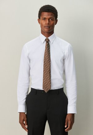 Mann trägt ein weißes Hemd, eine braune gemusterte Krawatte und schwarze Hose und steht vor einem schlichten hellen Hintergrund.