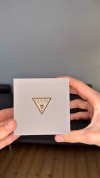 Caixa da marca Guess, em forma quadrada, com material cinza claro texturizado e um logótipo triangular dourado com um ponto de interrogação. Mãos visíveis a segurá-la.