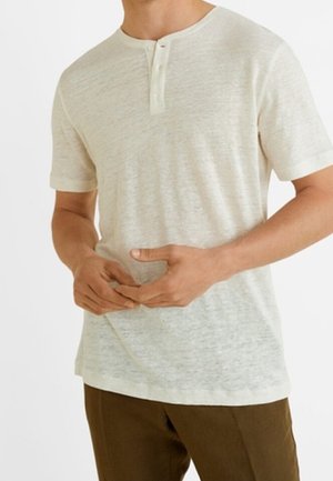 Mężczyzna w jasnobeżowej koszulce henley z krótkim rękawem i dwoma guzikami, w brązowych spodniach, z rękami splecionymi na wysokości pasa.