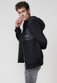 Chaqueta negra con cremallera y tejido texturizado, equipada con capucha. Presenta un panel negro contrastante con cremallera y detalle impreso. Combinada con pantalones gris claro.