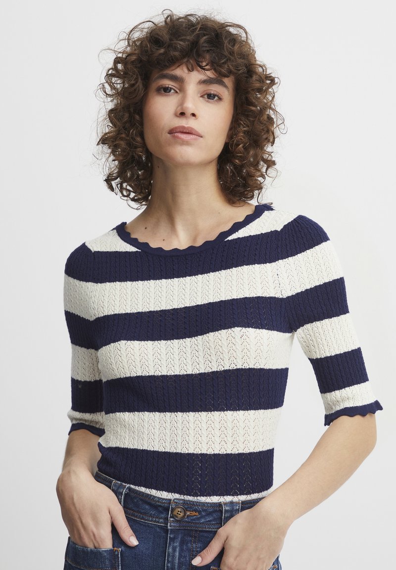Top in maglia a maniche corte con righe blu navy e bianche. Presenta un collo a onde e un tessuto texturizzato. Indossato con jeans a vita alta.