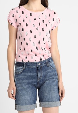 Nainen, jolla on pinkki lyhythihainen paita, jossa on mustavalkoinen jäätelökuvio ja käännetyt siniset farkkushortsit.