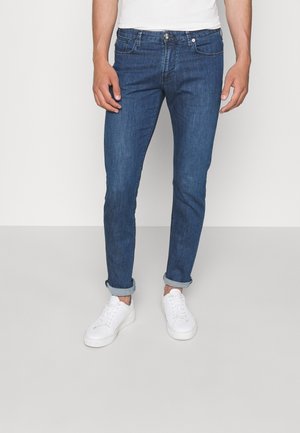 Mężczyzna ubrany w podwinięte niebieskie slim-fit jeansy, białe sneakersy i białą koszulę stoi na tle jednolitego jasnego tła.