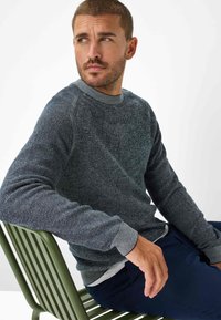 Struktureret mørkeblå sweater med rund hals og kontrasterende lys grå mansjetter. Båret over en hvid skjorte, siddende på en grøn metalstol.