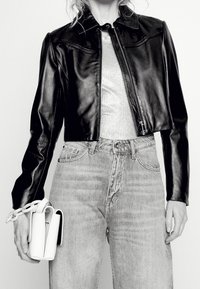 Chaqueta de cuero negra corta con un acabado brillante, cremallera frontal y cuello puntiagudo, combinada con jeans de tiro alto de lavado claro y un bolso blanco.