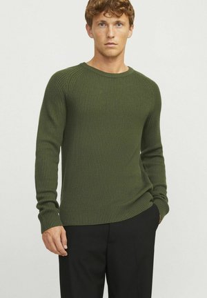 Maglione a coste di colore verde oliva scuro, con scollo rotondo, maniche lunghe e polsini aderenti. Indossato con pantaloni neri su misura.