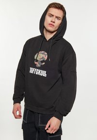 Fekete kapucnis pulóver kamuflázs koponya grafikával és "TUFFSKULL" felirattal. Állítható kapucnival, bordás mandzsettákkal és laza fazonnal készült.