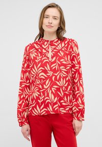 Blouse rouge avec un motif de feuilles crème sur l'ensemble, décolleté en keyhole avec quincaillerie, et longues manches froncées. Texture de tissu lisse.