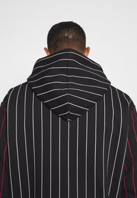 Musta huppari, jossa valkoiset pystysuorat raidat, muotoiltu huppu ja punaiset korostukset sivuilla. Pehmeää kangasta ja sileä pinta.