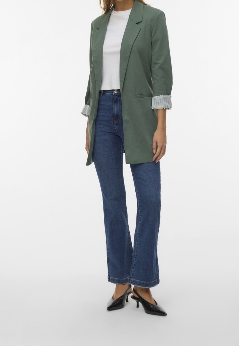Femme portant un blazer vert aux manches retroussées, un haut blanc court, un jean bleu taille haute, et des talons slingback noirs à bout pointu.