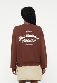 Sweatshirt castanho com gola redonda, apresentando texto branco "New Balance Athletics" e "Authentic" nas costas; punhos e bainha canelados.