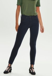 Vaqueros de mezclilla oscuros de cintura alta con un corte ajustado y un diseño clásico de cinco bolsillos. Llevados con una blusa verde con acentos de encaje.