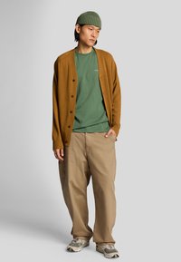 Brun strikket cardigan over en grøn t-shirt, parret med beige brede bukser og mønstrede sneakers. Modellen bærer en grøn ribbet hue.