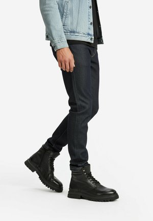 Person trägt dunkelblaue Slim-Fit-Jeans, schwarze Leder-Schnürstiefel und eine hellblaue Jeansjacke, steht vor einem weißen Hintergrund.