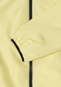 Manche de veste jaune clair avec poignet noir élastique et fermeture éclair noire verticale courant à côté de la manche sur le corps de la veste.