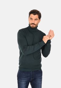 Maglione a collo alto di un verde scuro, realizzato in materiale morbido, design aderente con polsini e orlo a coste, abbinato a jeans blu scuro.