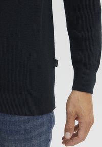 Pulover din tricot cu jaquard în nuanță bleu închis, cu un finisaj texturat, având un etichetă pe lateral și manșete fitted, purtat peste blugi denim de culoare bleu deschis.