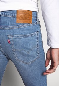 Světle modré džíny s tapered střihem, s béžovou koženou nášivkou na zadní straně, červenou etikettou na kapse a jemnými švy.