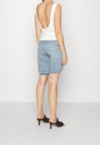 Haut sans manches blanc avec un dos dégagé, associé à un short en denim bleu clair aux bords effilochés, complété par des mules à talons noirs.