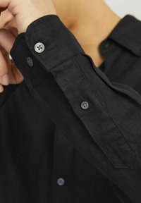 Camicia in lino nera con polsini abbottonati e una texture liscia. Il tessuto presenta una leggera lucentezza, con bottoni neri che rifiniscono i polsini e la patta.