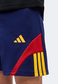 Σκούρο μπλε αθλητικό σορτς με κόκκινο κυρτό πάνελ και τρεις κάθετες κίτρινες ρίγες· κίτρινο λογότυπο Adidas στο αριστερό μηρό.