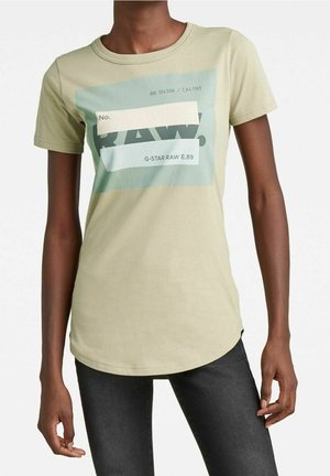 Slim-fit beige T-shirt met grafische "RAW" tekst en codes, gedragen door een persoon met donkere spijkerbroek, staand tegen een effen achtergrond.