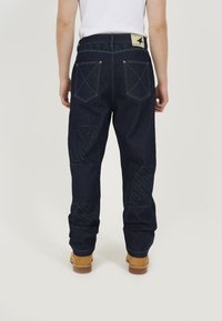 Dark blue denim pants with green stitching, featuring large back pockets and logo patterns on the side. Flat waistband and straight legs.