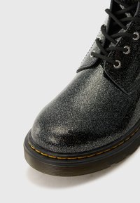 Botín negro de purpurina de cuero con punta redonda, cordones negros, ojetes circulares y una suela amarilla cosida para contraste.