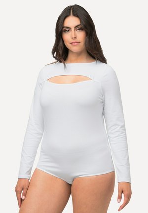 Mujer con cabello largo y oscuro que viste un bodysuit blanco de manga larga con un corte horizontal por encima del pecho, de pie contra un fondo liso.