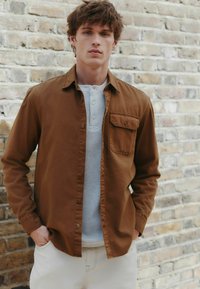 Hnedá, dlhá košeľa s gombíkmi cez svetlomodrý henley, doplnená krémovými nohavicami. Viditeľná textúrovaná tkanina a detail prednej vrecka.