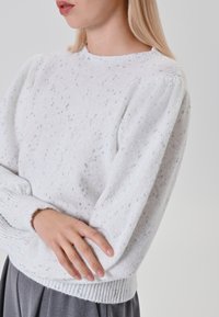 Maglione bianco lavorato a maglia con glitter argentati, scollatura rotonda, maniche lunghe a sbuffo e polsini e orlo a costine. Indossato con una gonna grigia.