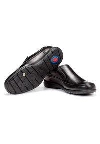 Zapatos de cuero negro tipo slip-on con un upper liso, suela acolchada y suela exterior de goma texturizada que presenta un logo rojo y azul en el talón.