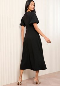 Zwart midi-jurk met pofmouwen en een aansluitend lijfje. Glad materiaal met zij-splitdetail. Model draagt doorzichtige hakken.