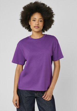 Femme aux cheveux bouclés naturels portant un t-shirt violet uni à manches courtes et un jean foncé, debout devant un fond gris clair.