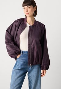 Giacca bomber imbottita viola con zip frontale, colletto a costine e polsini elastici. Indossata su una maglietta aderente bianca e jeans azzurri chiari.