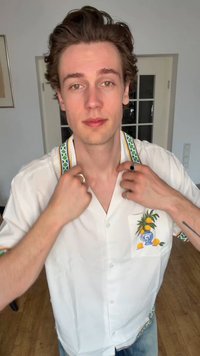 Νέος άνδρας ρυθμίζει τον γιακά ενός λευκού κοντομάνικου πουκάμισου με κέντημα λεμονιού και αγγείου, στέκεται σε εσωτερικό χώρο με ξύλινο πάτωμα και πόρτα με τζάμι πίσω του.