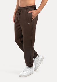 Braune Jogginghosen mit einem elastischen Bund, Seitentaschen und elastischen Knöcheln, kombiniert mit weißen Turnschuhen. Glatte, weiche Textur.