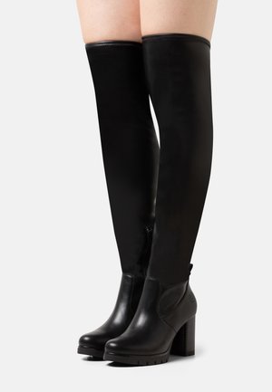 Femme portant des bottes noires montantes jusqu'aux cuisses avec des talons blocs et une fermeture éclair latérale sur un fond clair uni.