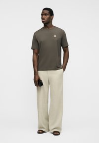 Férfi olívazöld pólóban, kis rókalogóval, bézs, széles szárú nadrágban, fekete szandálban, kezében egy fekete, összecsukható pénztárcával.