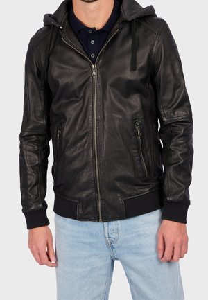 Mann trägt eine schwarze Lederjacke mit Reißverschluss und Kapuze, vorn mit Reißverschlusstaschen und schwarzen Bündchen, kombiniert mit hellblauen Jeans.