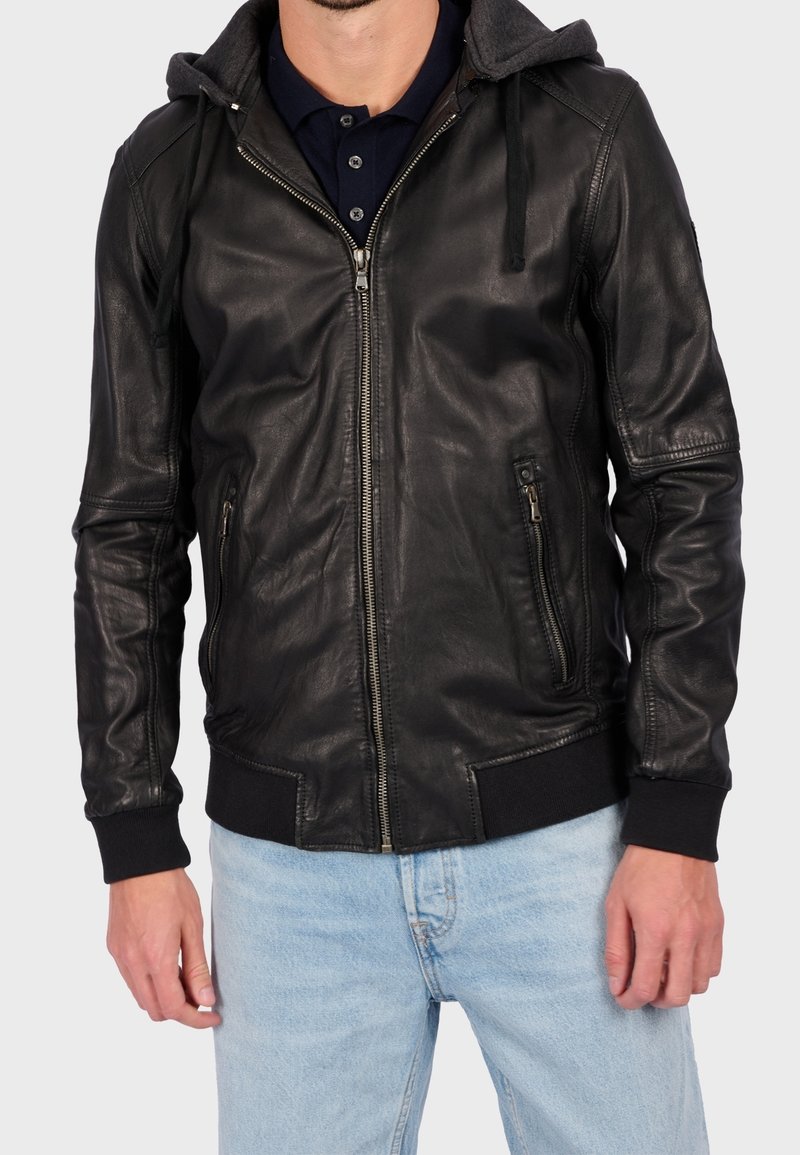Mężczyzna w czarnej skórzanej kurtce zapinanej na zamek z kapturem, przednimi kieszeniami na suwak i czarnymi ściągaczami, w połączeniu z jasnoniebieskimi dżinsami.