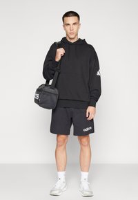Junger Mann trägt schwarzen Adidas-Hoodie und Shorts, weiße Sneaker, trägt schwarze Adidas-Tasche über der Schulter, steht auf grauem Boden.