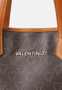 Ruda tekstūruota pirkinių rankinė su šviesiai rusvais odiniais rankenomis. Auksinė "VALENTINO" logotipas priekyje. Medžiagoje yra subtilus reljefinis raštas.