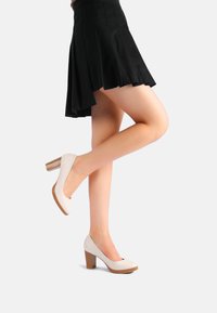 Scarpe con tacco alto bianche, con punta arrotondata e tacco impilato marrone testurizzato. Indossate con una gonna nera a vita alta, che mette in mostra la gamba.