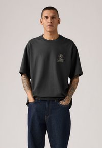 Άνδρας με κοντά μαλλιά, φοράει χαλαρό σκουρόχρωμο γκρι μπλουζάκι με μικρό λουλουδάτο σχέδιο και σκουρό μπλε τζιν, με τα χέρια στις τσέπες, στέκεται μπροστά σε απλό φόντο.